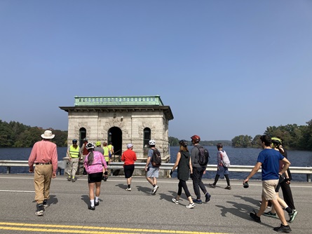 People walking across a road to enter the Hobbs Brook Reservoir gatehouse - a gray stone building. Blue sky and the reservoir are seen in the background.