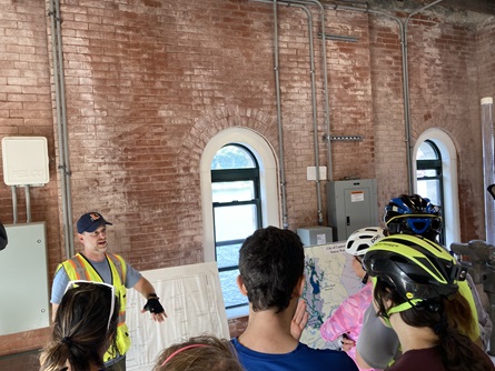 A staff member gives a talk using diagrams inside the Stony Brook Reservoir Gatehouse. People stand in front of the red brick walls.