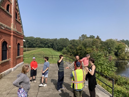 Staff members and participants gather on the Stony Brook Dam to chat. The red brick gatehouse and grassy dam are seen in the background.