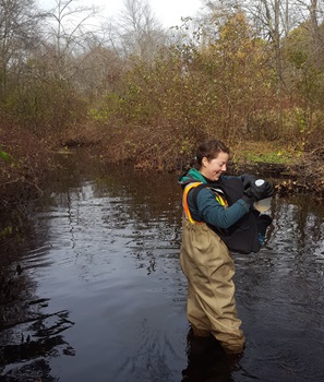 A person stands in stream holding a water quality sampling bottle