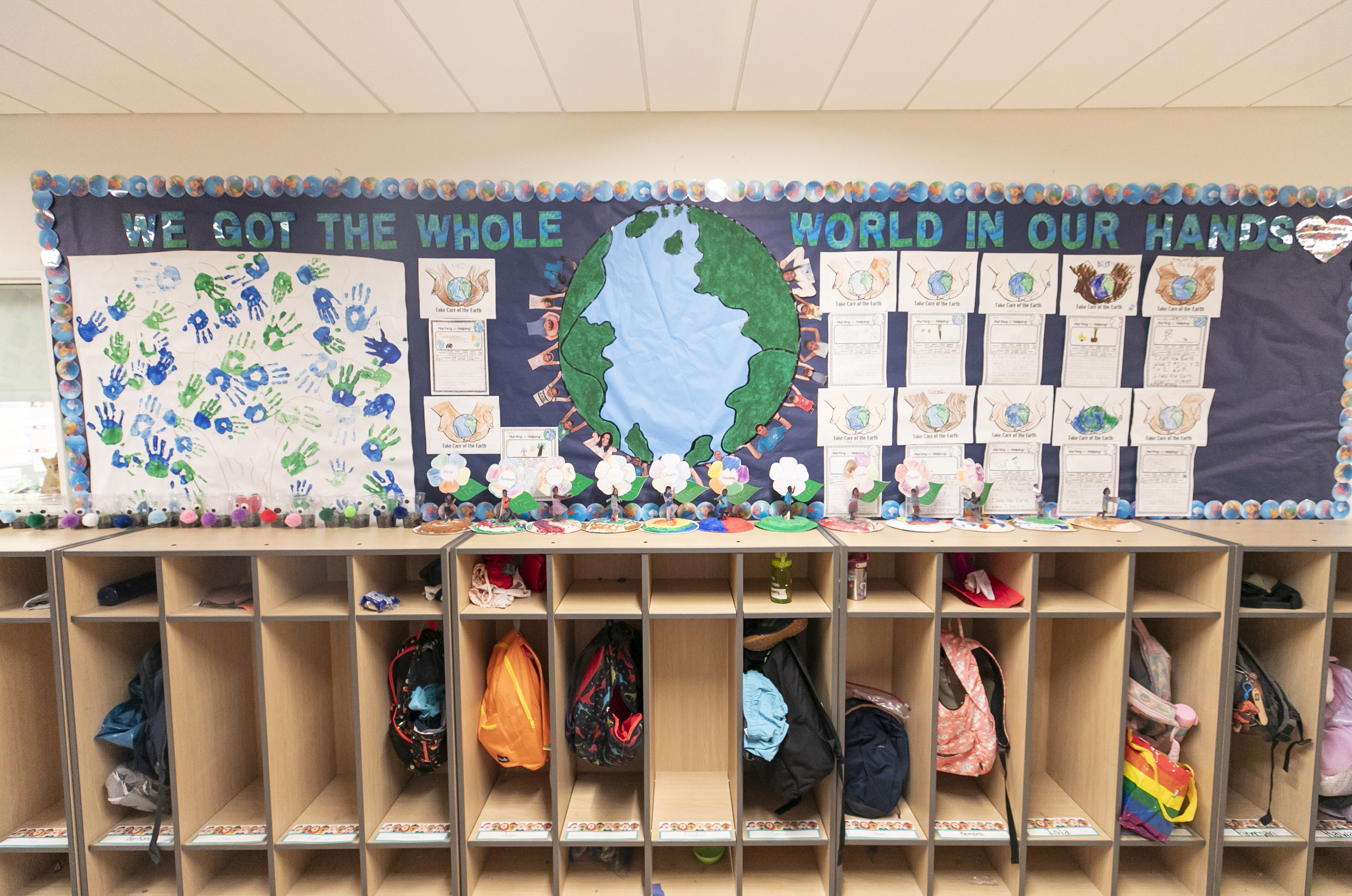 Children's backpacks in cubby storage. A bulletin board with handprints is above it