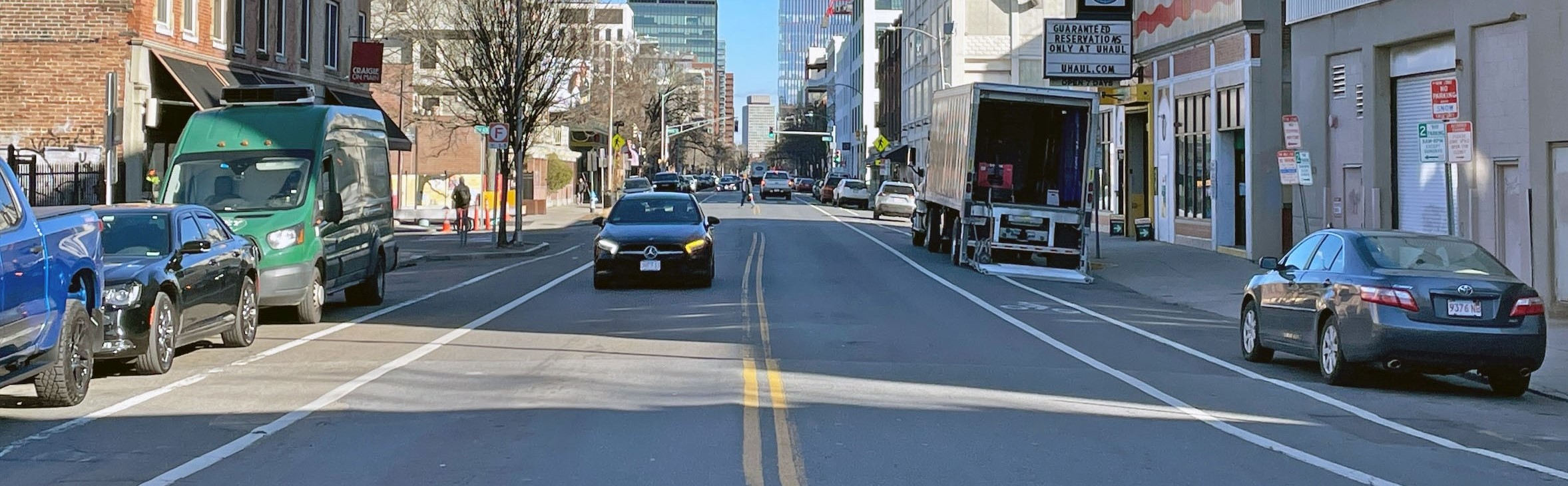 A view of Main Street in Cambridge shows a UHaul storefront with a moving truck parked out front, several cars on the road, and a pedestrian crossing in the background. Tere are traditional bike lanes in both directions.