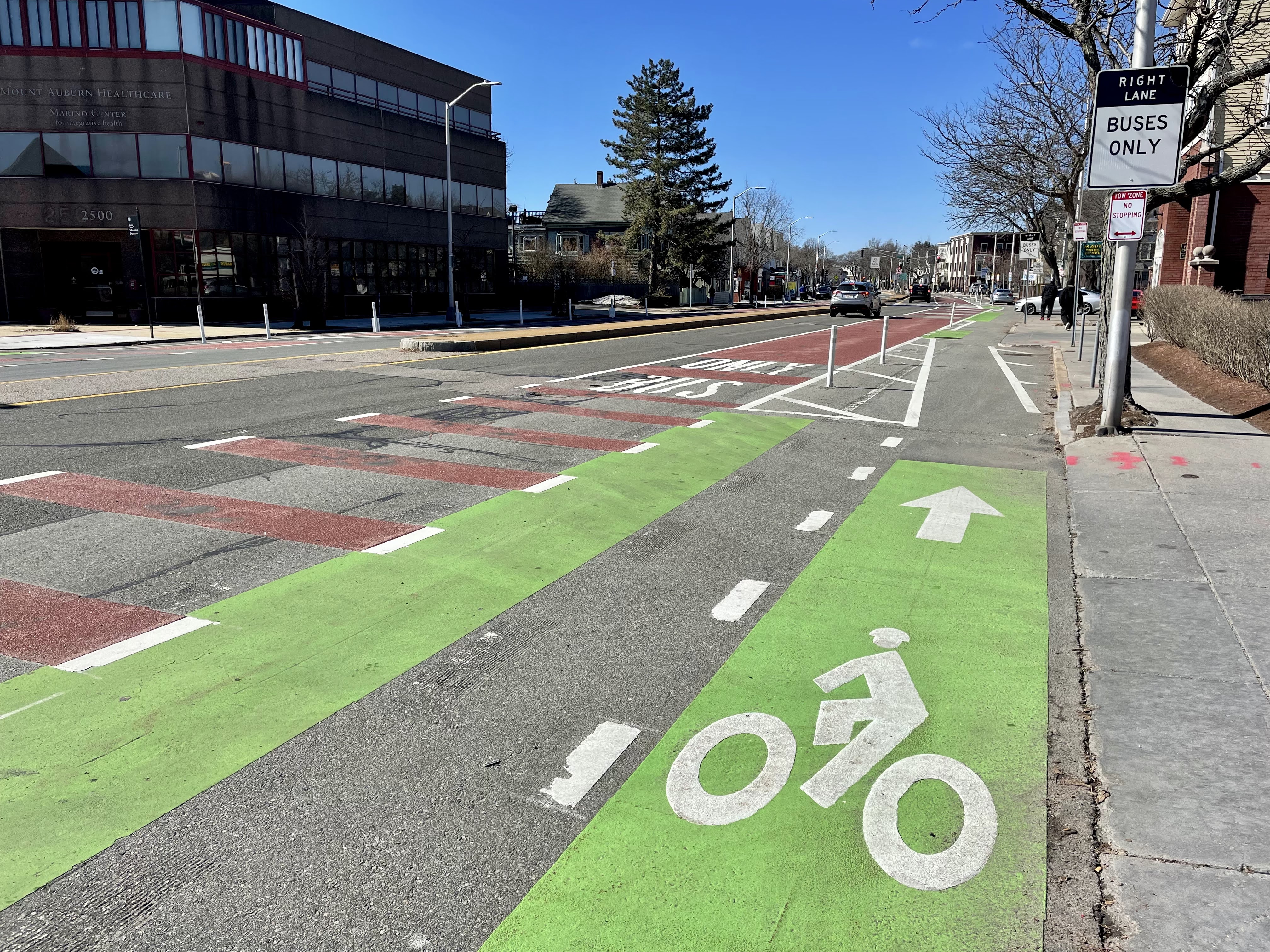 Image depicts a street - on the sidewalk is a sign that says "Right Lane Buses Only." Next to the sidewalk, a bike lane is next to the curb, separated from the rest of the street by flex posts. Nex tot the bike lane, there is a red travel lane with markings that say "Only Bus." Next to the bus lane is a travel lane with several vehicles, separated from the other side of the street with a median.