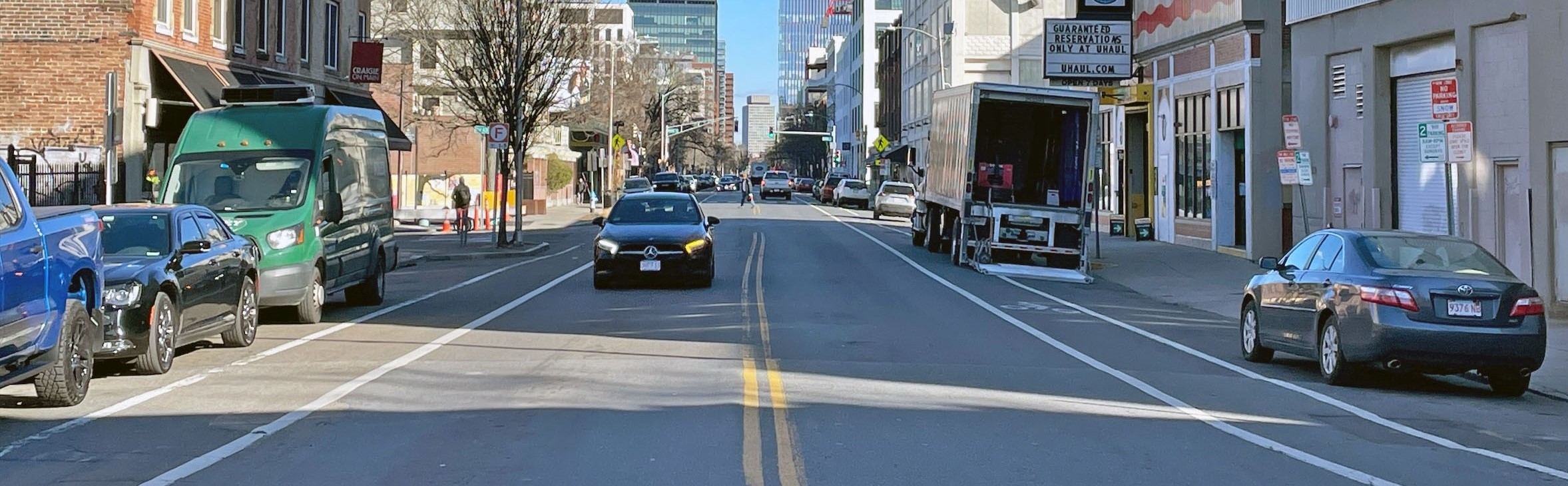 A view of Main Street in Cambridge shows a UHaul storefront with a moving truck parked out front, several cars on the road, and a pedestrian crossing in the background. Tere are traditional bike lanes in both directions.