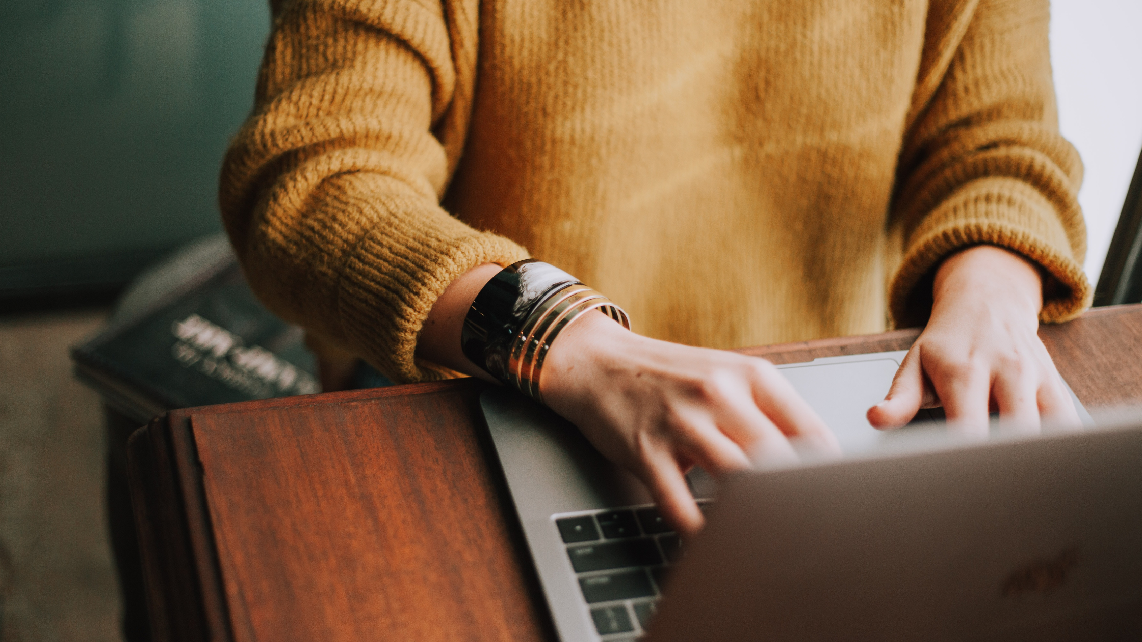 Person in yellow sweater typing on a silver laptop on a wooden table.