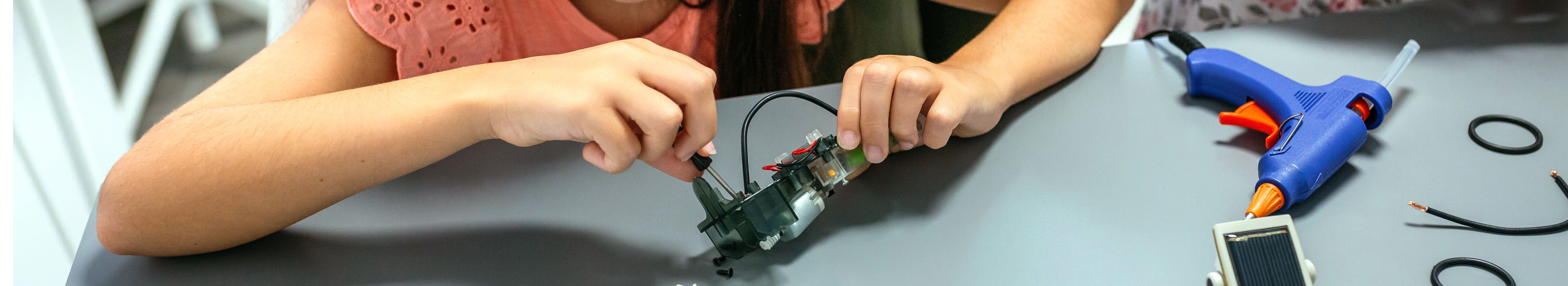 A child uses a screwdriver to tighten a piece of electronic gear, with a hot glue gun lying on the table nearby