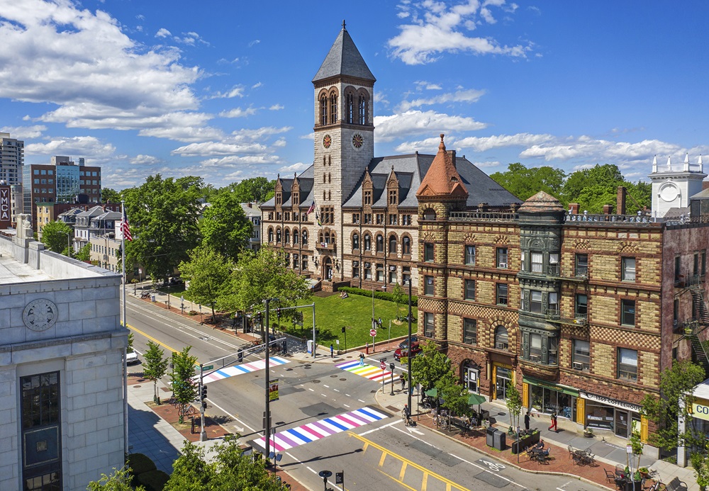 City Hall stands tall on a bright, sunny day with blue skies and a few clouds. Neighboring buildings are shown, as well as pedestrians, bikers, and cars on Mass Ave, and there are colorful crosswalks painted with Pride colors.