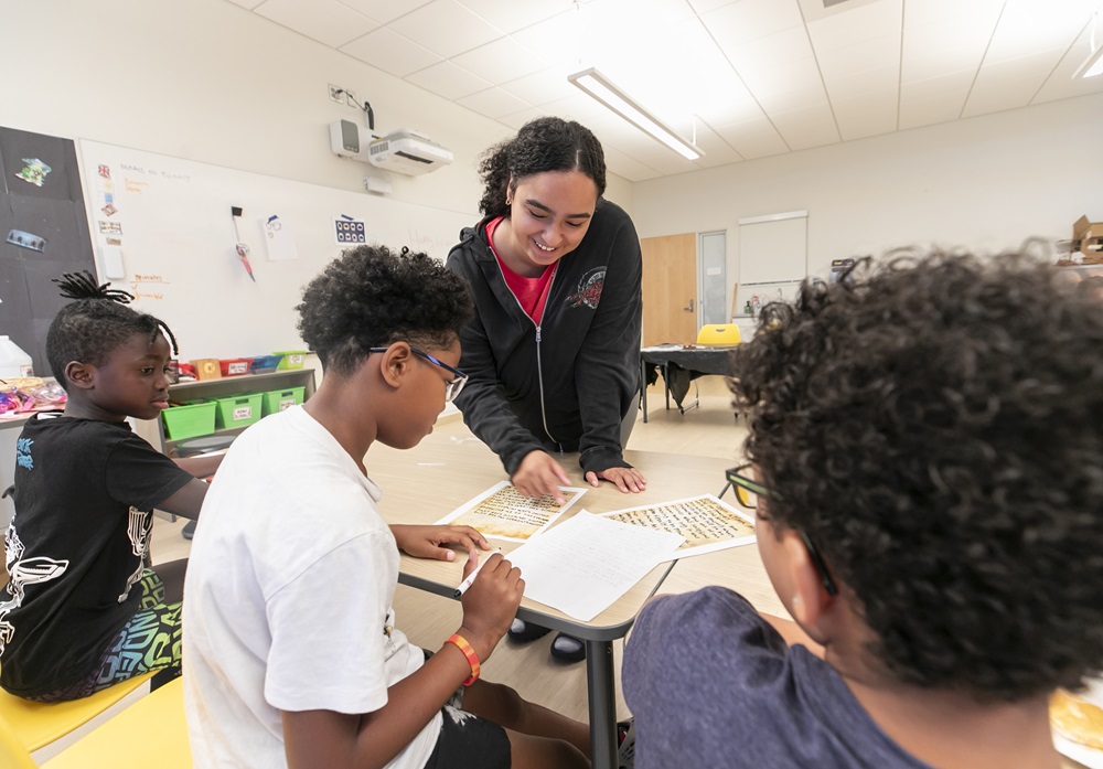 A smiling teacher supervises three students working on a classroom assignment at a table, pointing at one of their papers. The room is bright with educational posters visible in the background.