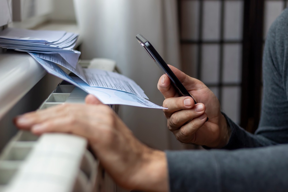 A person reviews energy bills while holding a phone next to a window.