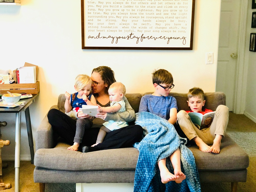 A family of five, a mother and four children, sit closely together on a sofa, engaged in reading books. The room appears cozy, illuminated by natural light.