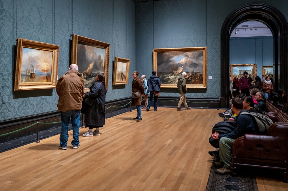 Visitors observing paintings, with some seated on brown leather benches and others standing close to the artworks.