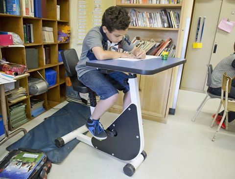 Boy sitting on a bicycle desk in his classroom doing school work