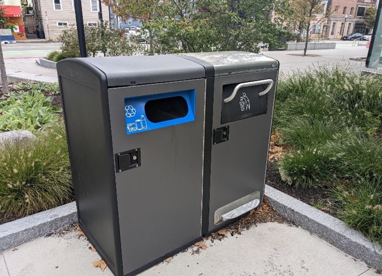 An enclosed trash can with a foot peddle next to a recycling bin.